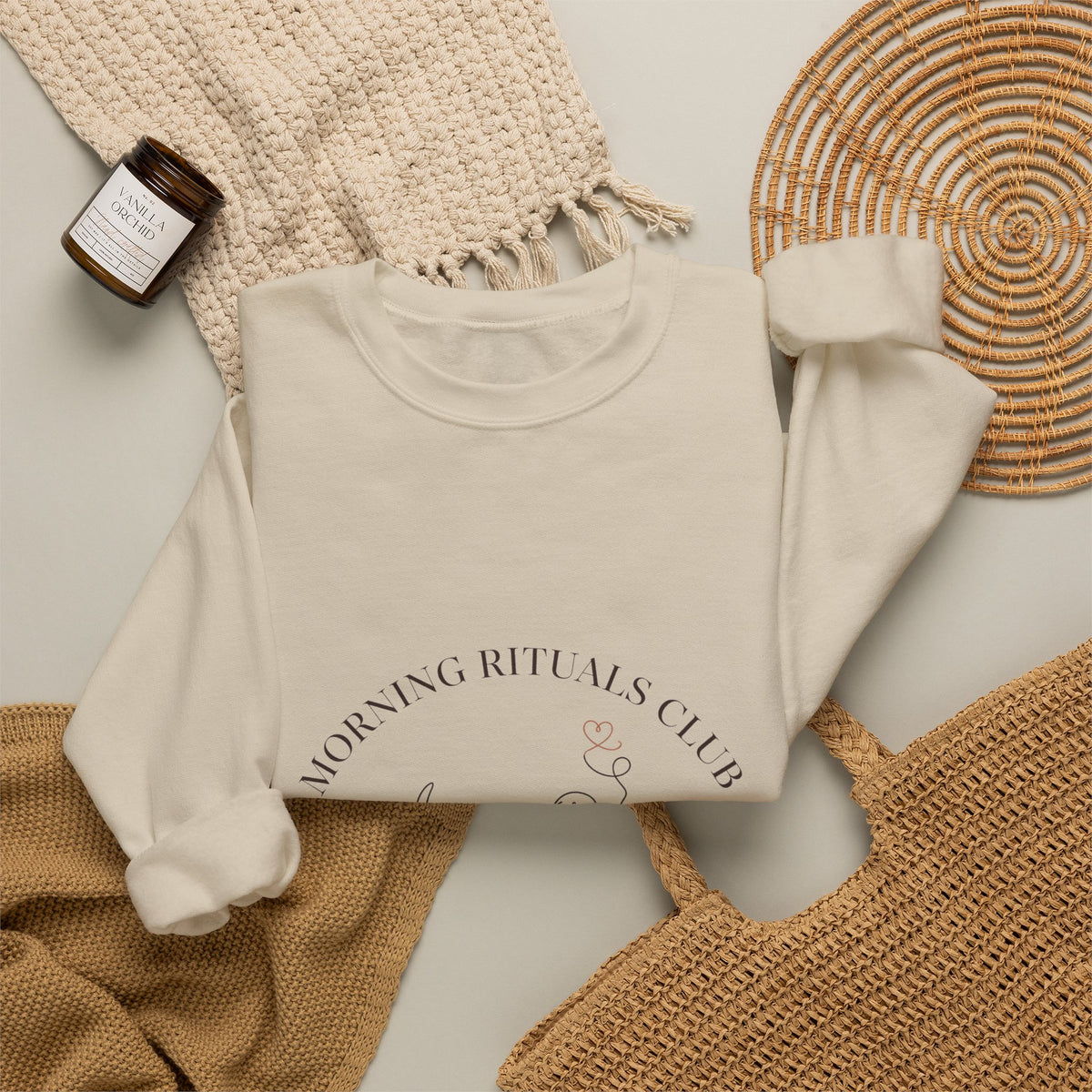 Morning Rituals Club crewneck folded flat with a candle, woven placemat, and knit throw