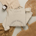 Sunday Morning Sofa Club crewneck folded flat with a candle, woven placemat, and knit throw