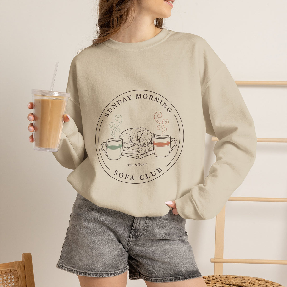 Woman wearing the sand Sunday Morning Sofa Club crewneck while holding an iced coffee